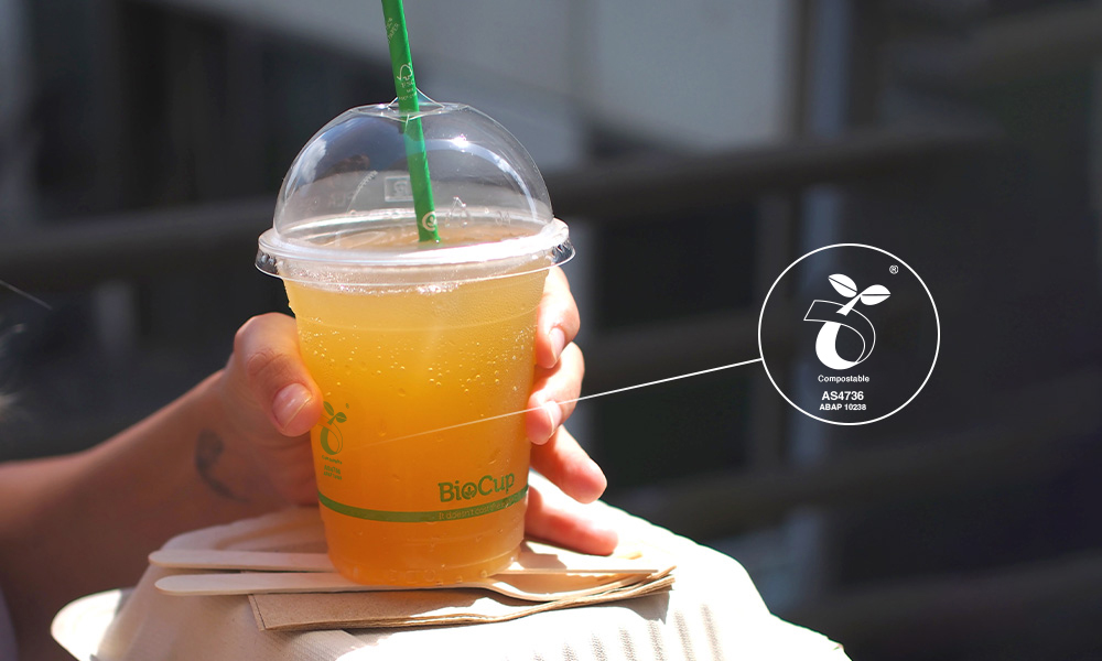 A hand holding up PLA bioplastic BioCup with orange juice. The image zooms in on the compostable label, reading ABAP10238.