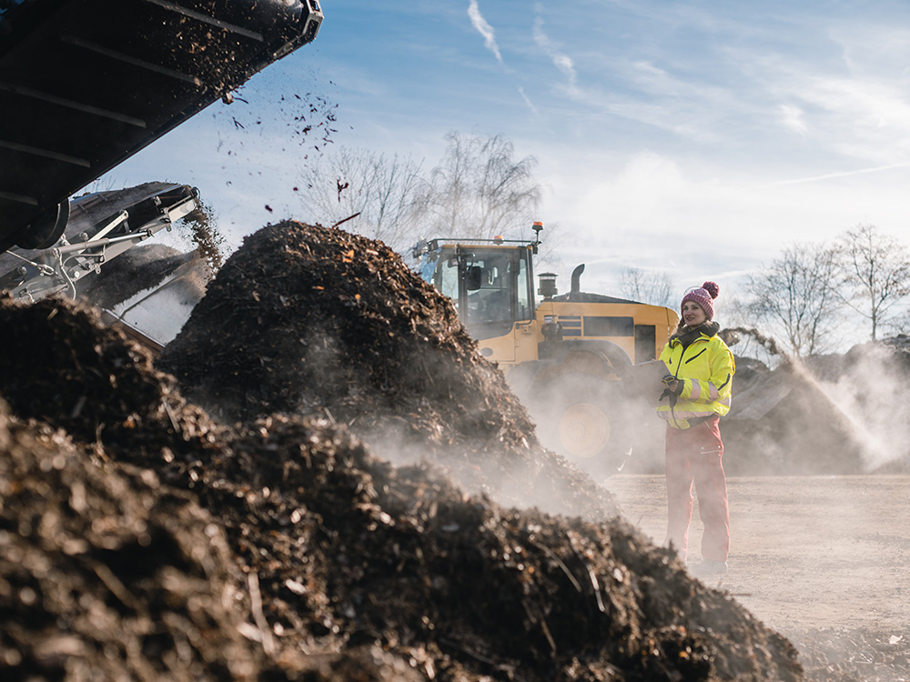 An industrial composting facility with a pile of organic matter and compost. There’s heavy machinery in the background moving the compost around.
