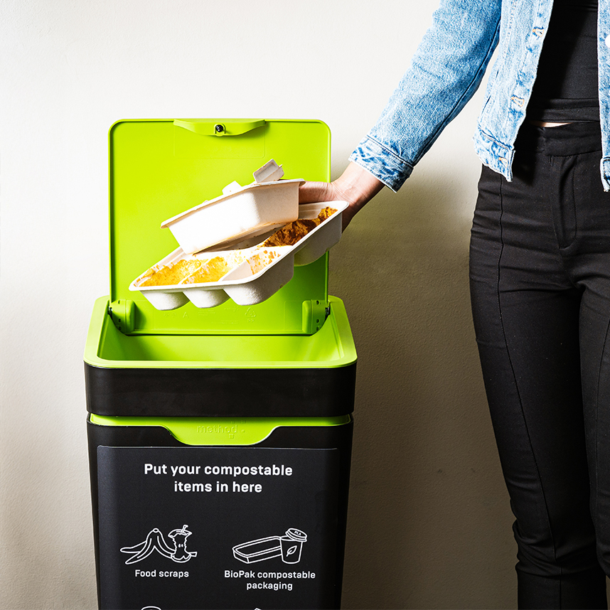 Person disposing compostable trays into a green-lid compost bin.